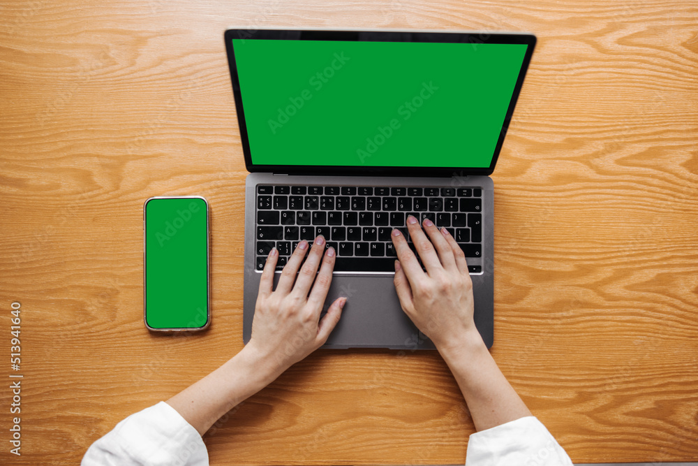 Fototapeta premium Young caucasian woman work at office desk using laptop and smartphone. Blank green screen of wireless devices. Technology concept
