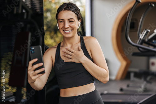 Happy young caucasian woman smiling video call via phone in sport club. Brunette wears black top and leggings in training. Technology concept