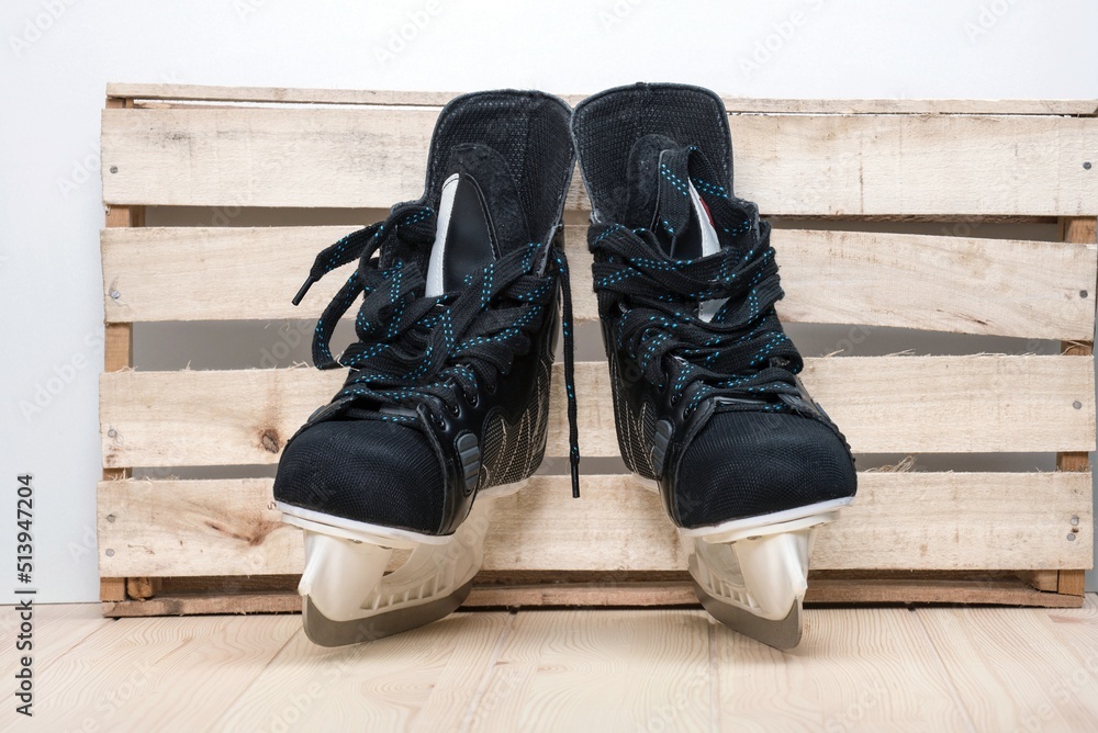 A pair of black hockey skates with well-sharpened metal blades in a white plastic frame designed to play ice hockey as a popular winter sport stay next to the craft wooden wall waiting for game starts