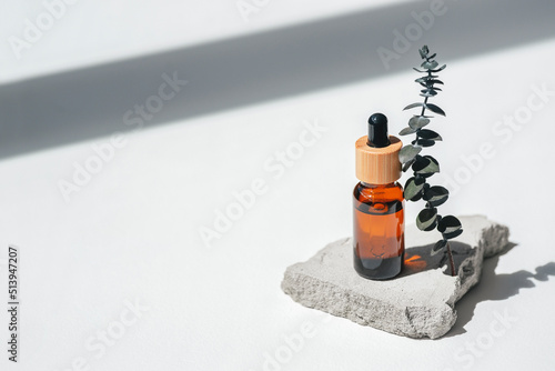 Amber bottle with dropper pipette and serum or essential oil on grey concrete podium with eucalyptus branches. White background with daylight. Beauty concept for face and body care