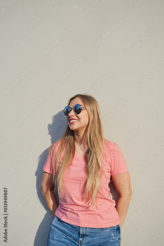 Happy, smiling blonde woman in sunglasses, in a pink t-shirt, standing against a gray wall on a sunny day Happy, smiling blonde woman in sunglasses, in a pink t-shirt, standing against a gray wall on a sunny day