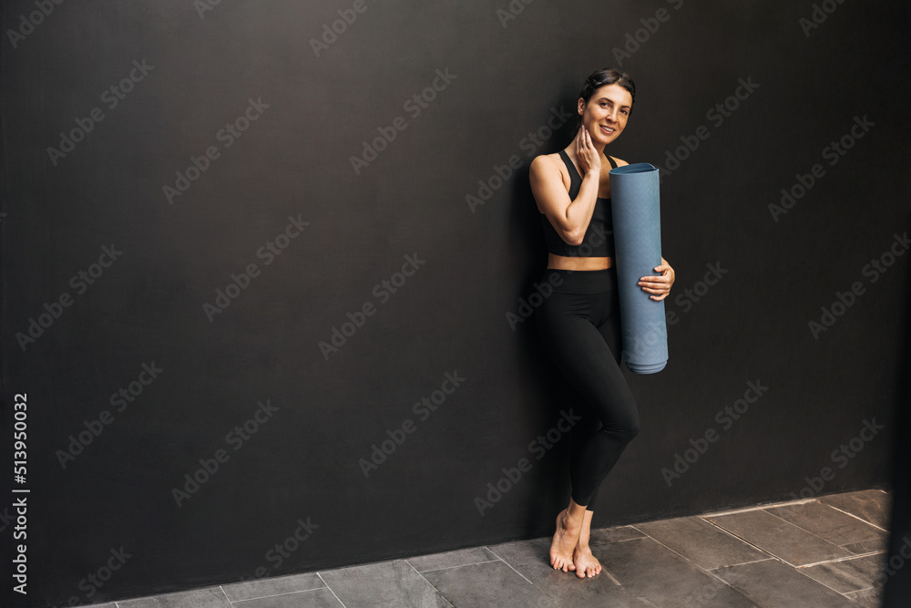 Full length young caucasian girl with slim body shape holds mat standing near wall with space for text. Brunette wears black sports top and leggings. Healthy lifestyle concept Full length young caucasian girl with slim body shape holds mat standing near wall with space for text. Brunette wears black sports top and leggings. Healthy lifestyle concept