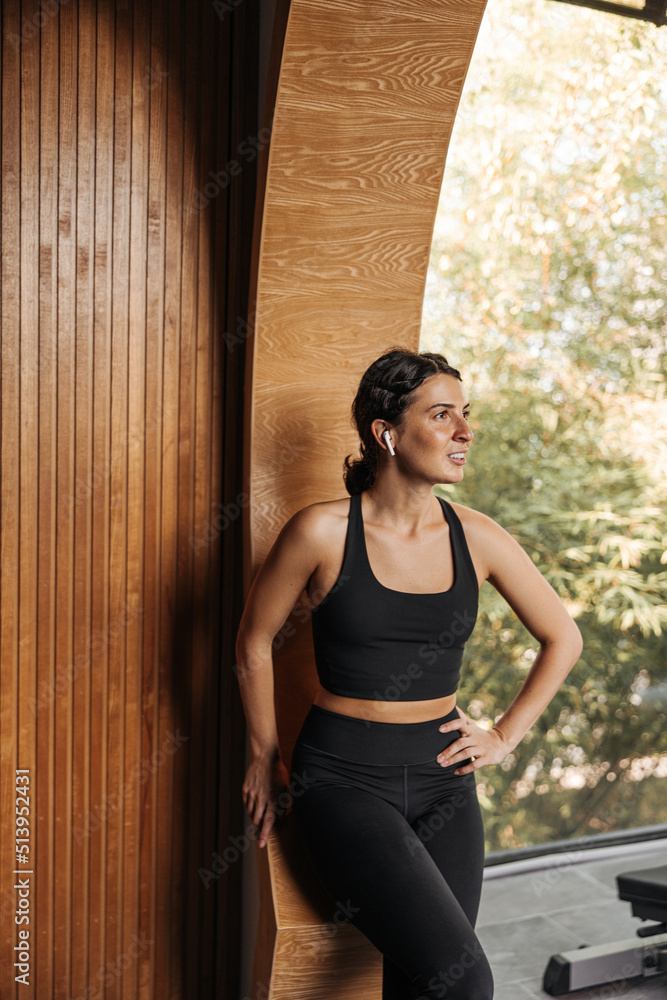 Athletic young caucasian woman in sportswear looks away standing in gym. Brunette wears black top and leggings in training. Healthy lifestyle concept.