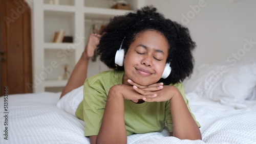 Funny woman lies on the bed in the room listens to music on headphones and dances. Mixing races results in beautiful people. The routine life of Americans in their free time.