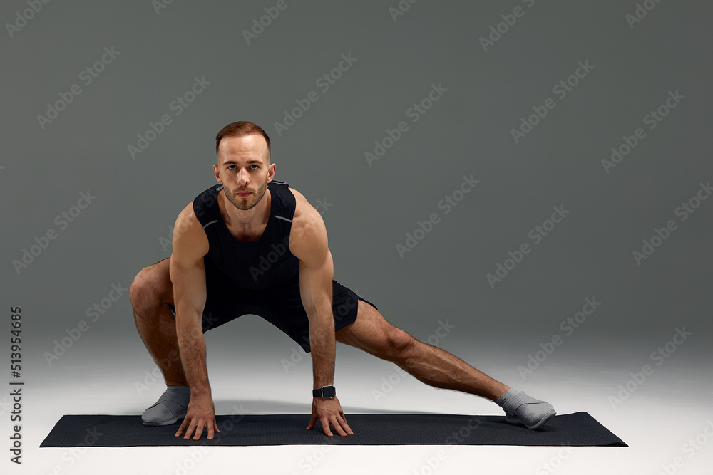 Attractive healthy young sports man stretching on fitness mat indoors, over gray wall background