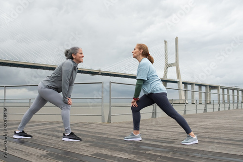 Dynamic senior friends exercising on embankment. Women in sportive clothes stretching on cloudy day. Sport, friendship concept
