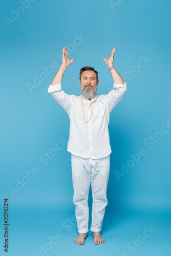 full length of yoga master meditating with raised hands white standing on blue.