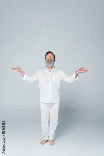 full length of smiling yoga guru meditating in tree pose with open arms on grey.