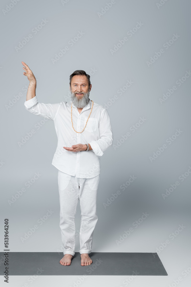 bearded and barefoot guru man in white clothes gesturing and looking at camera on grey. bearded and barefoot guru man in white clothes gesturing and looking at camera on grey.