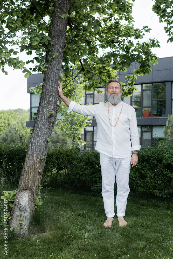 full length of guru coach touching tree trunk while meditating with closed eyes outdoors. full length of guru coach touching tree trunk while meditating with closed eyes outdoors.