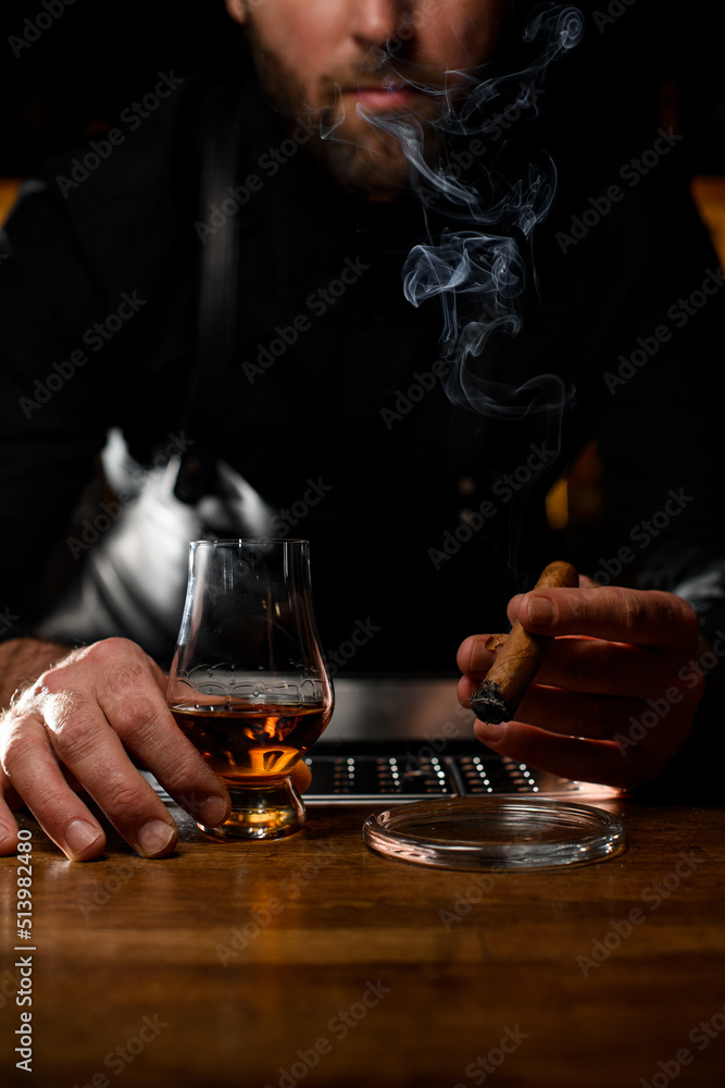 view on male hand holding glass with cognac and smoking cuban cigar over ashtray