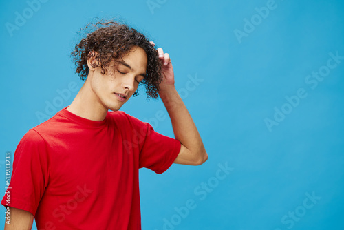 Pensive thoughtful unhappy young curly tanned Caucasian guy in basic red t-shirt posing isolated on over blue background. Lifestyle and Emotions concept. Good offer with copy space for ad
