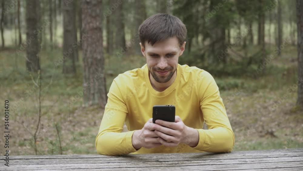 Caucasian man in yellow longsleeve is sitting at wooden table in forest and typing text message on smartphone with smile, playing mobile game. Positive brunette uses phone, looks at screen in nature. Caucasian man in yellow longsleeve is sitting at wooden table in forest and typing text message on smartphone with smile, playing mobile game. Positive brunette uses phone, looks at screen in nature.