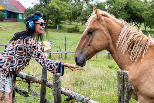A beautiful girl in glasses and with a bandana on her head feeds the horses in the meadow. Blurred background.