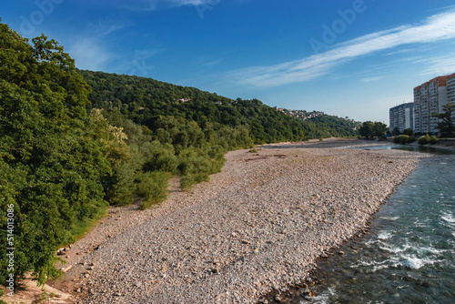 A shallow river flows in the city limits against the background of a bank covered with dense green vegetation. View of the mountain river on a sunny summer day under a blue sky with white clouds. © Vit-Vit