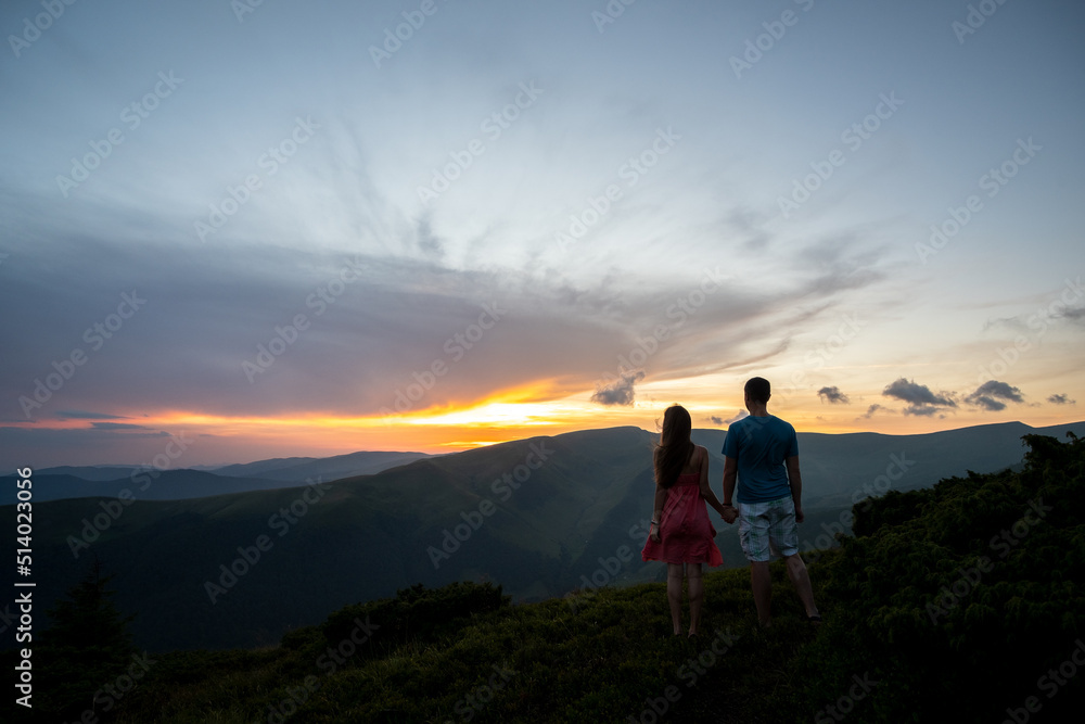 Back view of young hiker couple standing holding hands on grassy mountain hill enjoying sunset panorama and dramatic evening sky