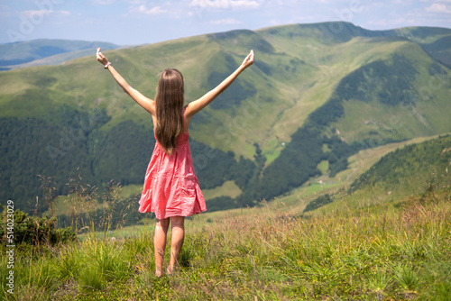 Back view of young happy woman traveler in red dress standing on grassy hillside on a windy day in summer mountains with outstretched arms enjoying view of nature