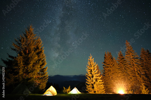 Fényképezés Bright illuminated tourist tents near glowing bonfire on camping site in dark mountains under night sky with sparkling stars