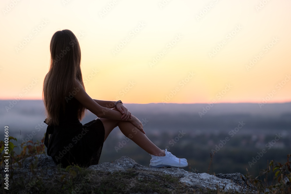 Young elegant woman in black short dress and white sneaker shoes sitting on a rock relaxing outdoors at summer evening. Fashionable lady enjoying warm sunset in nature