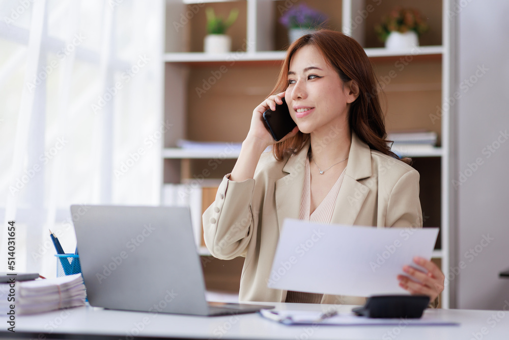 Portrait of a young Asian woman sitting at an office desk with a laptop computer and files and talking on a mobile phone. Communication concept Portrait of a young Asian woman sitting at an office desk with a laptop computer and files and talking on a mobile phone. Communication concept