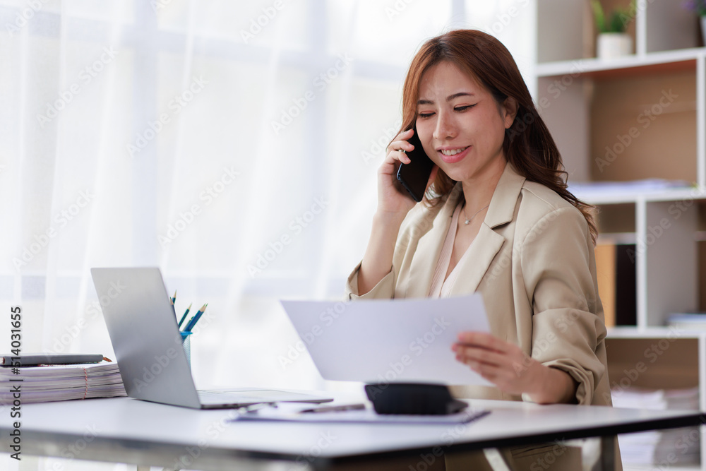 Portrait of a young Asian woman sitting at an office desk with a laptop computer and files and talking on a mobile phone. Communication concept Portrait of a young Asian woman sitting at an office desk with a laptop computer and files and talking on a mobile phone. Communication concept