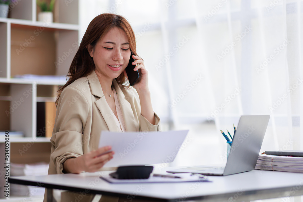 Portrait of a young Asian woman sitting at an office desk with a laptop computer and files and talking on a mobile phone. Communication concept Portrait of a young Asian woman sitting at an office desk with a laptop computer and files and talking on a mobile phone. Communication concept