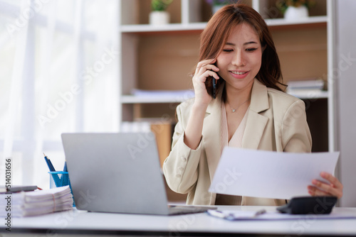 Portrait of a young Asian woman sitting at an office desk with a laptop computer and files and talking on a mobile phone. Communication concept