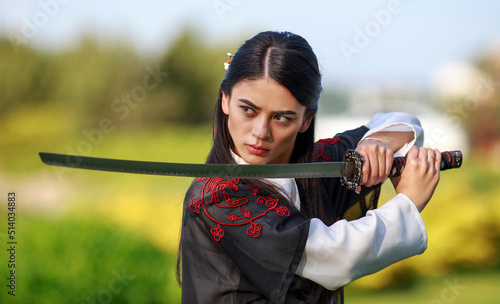 Young asian woman in traditional kimono trains in a fighting stance close-up portrait with katana sword samurai warrior girl in green summer garden