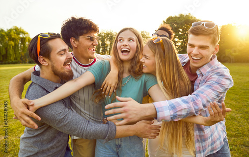 Group of funny, excited friends fooling around and having fun in the park. Bunch of happy, joyful, smiling people standing on a green lawn and all together hugging a cheerful beautiful young girl photo