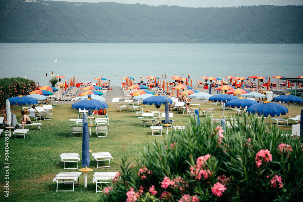 Lake beach with colorful umbrellas and lounge chairs on green lawn grass with pink oleander flowers bush at the foreground