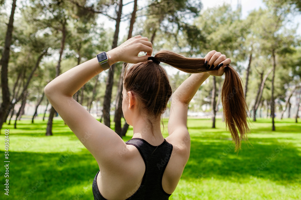 Fotografie Young brunette woman wearing sportive clothes on city park, outdoors tying her hair in a ponytail while doing some workout