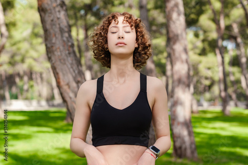 Portrait of redhead woman wearing black sports bra standing on city park, outdoors enjoying stress free mindful moment, doing yoga relaxation exercises, closed eyes, meditating nature, taking breath.