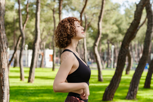 Side view of cute sportive woman wearing black sports bra standing on city park, outdoors enjoying stress free mindful moment, doing yoga relaxation exercises, closed eyes, meditating nature. photo