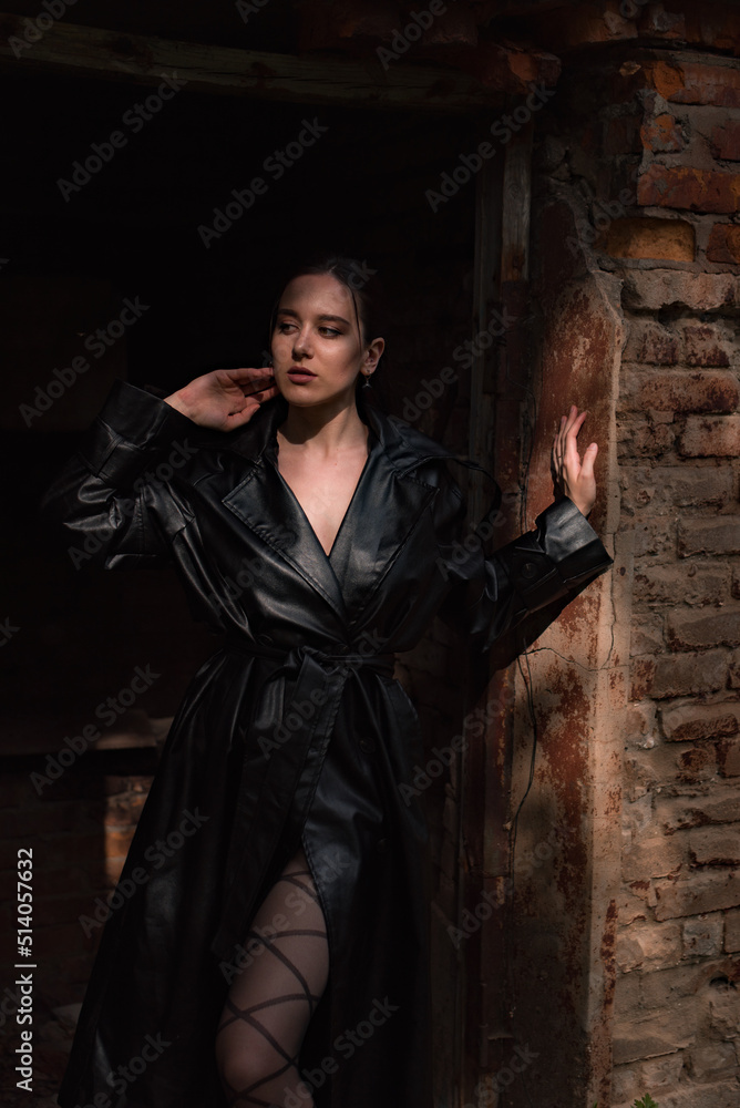 a young pretty woman in a black leather coat, raincoat in a park near a destroyed building a young pretty woman in a black leather coat, raincoat in a park near a destroyed building