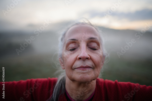 Senior woman doing breathing exercise in nature on early morning with fog and mountains in background, close-up © Halfpoint