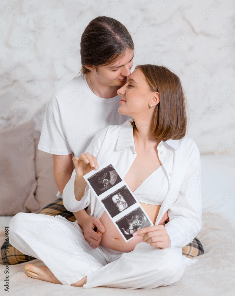 Front view of couple expecting baby, tender man kissing forehead behind of beautiful pregnant woman which sitting on bed in lotus pose and holding ultrasound monochrome image of womb with baby Photos 