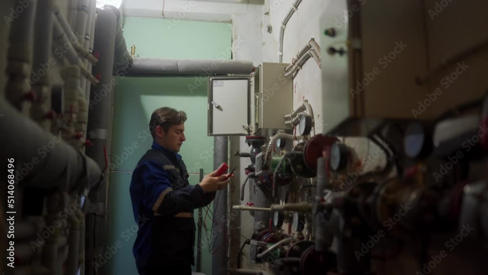 Electrician inspecting control panel with multimeter in a district heating substation. Master in uniform checking fuse box with tester probes to fix electricity. Man working in a technical room. Electrician inspecting control panel with multimeter in a district heating substation. Master in uniform checking fuse box with tester probes to fix electricity. Man working in a technical room.