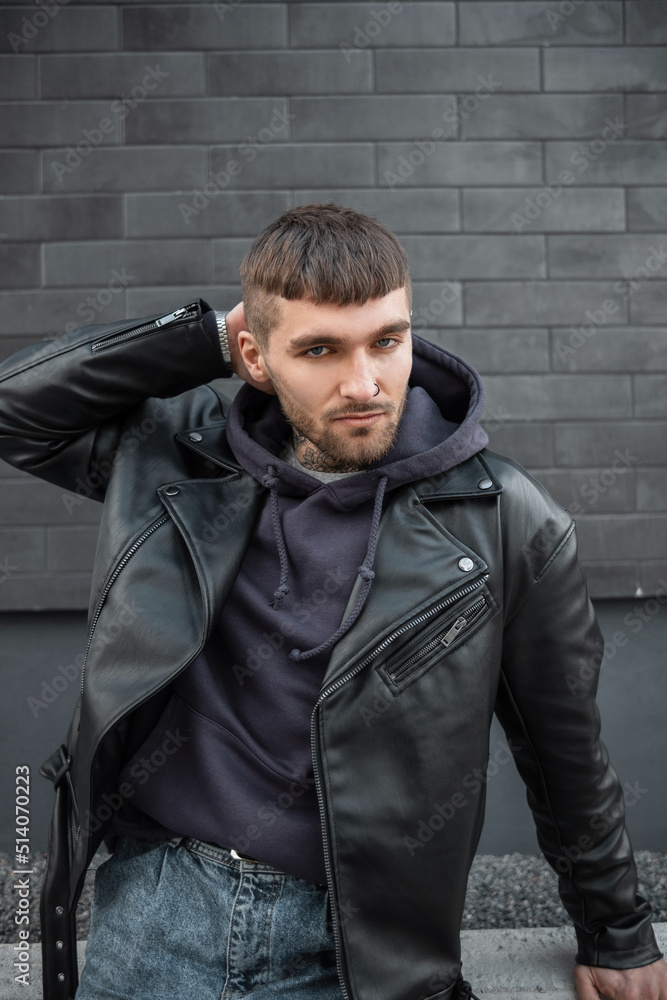 Fashionable hipster man with hairstyle in stylish casual outfit in leather jacket, hoodie and jeans sits and poses near a black brick wall on the street
