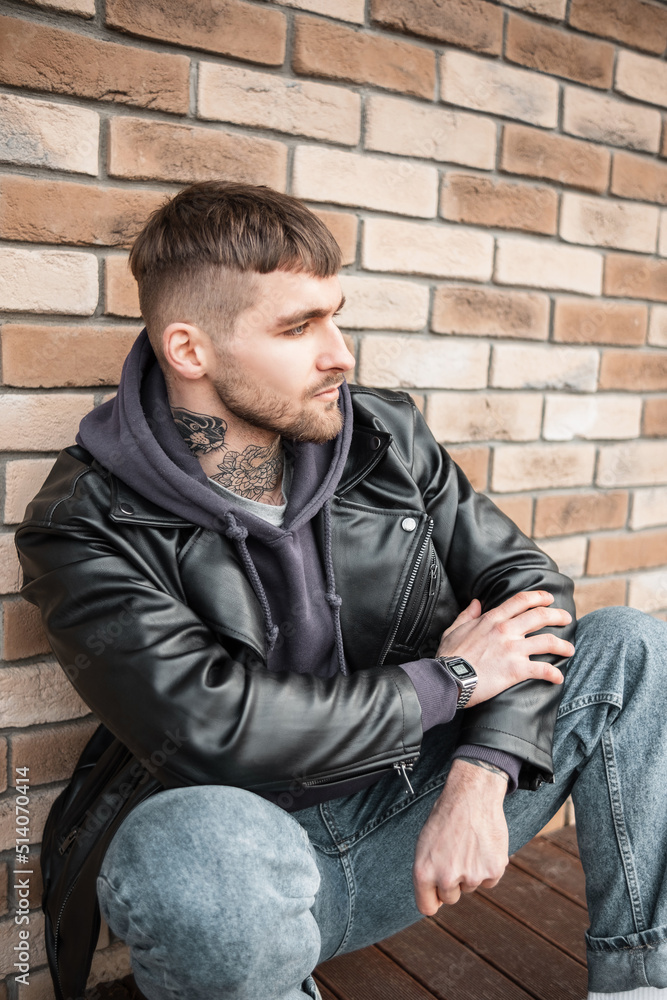Stylish handsome American model hipster guy with hair and tattoos on his neck in fashionable casual clothes sits near a brick wall