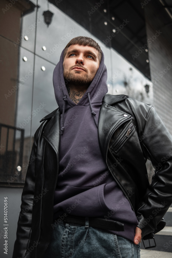 Obraz premium Fashionable handsome man model hipster in stylish clothes with a rock fashion black jacket and hoodie walking in the city