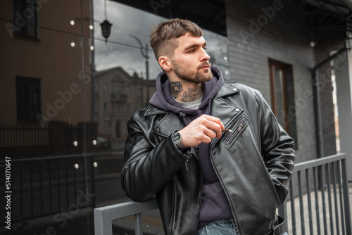 Handsome brutal hipster guy with a cigarette in fashion black outfit with leather jacket and hoodie walking in the city and smoking a cigarette photo