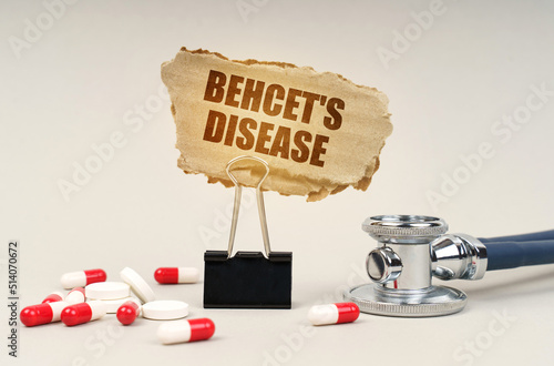 Near the stethoscope are pills and a clip with a cardboard sign - Behcets disease