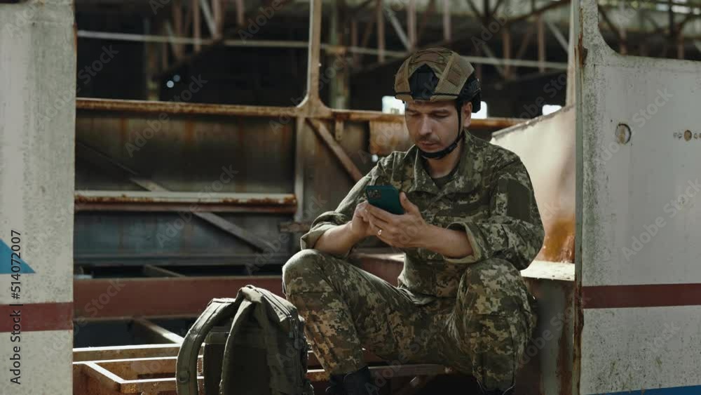 Happy man in military uniform scrolling social media on modern smartphone while sitting inside abandoned building after war. Concept of armed forces and technology.