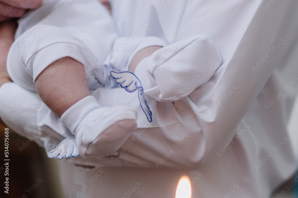 Fototapeta premium Infant baptism ceremony. Close up of tiny baby legs, sacrament of baptism. The godfather holds the child in his arms.