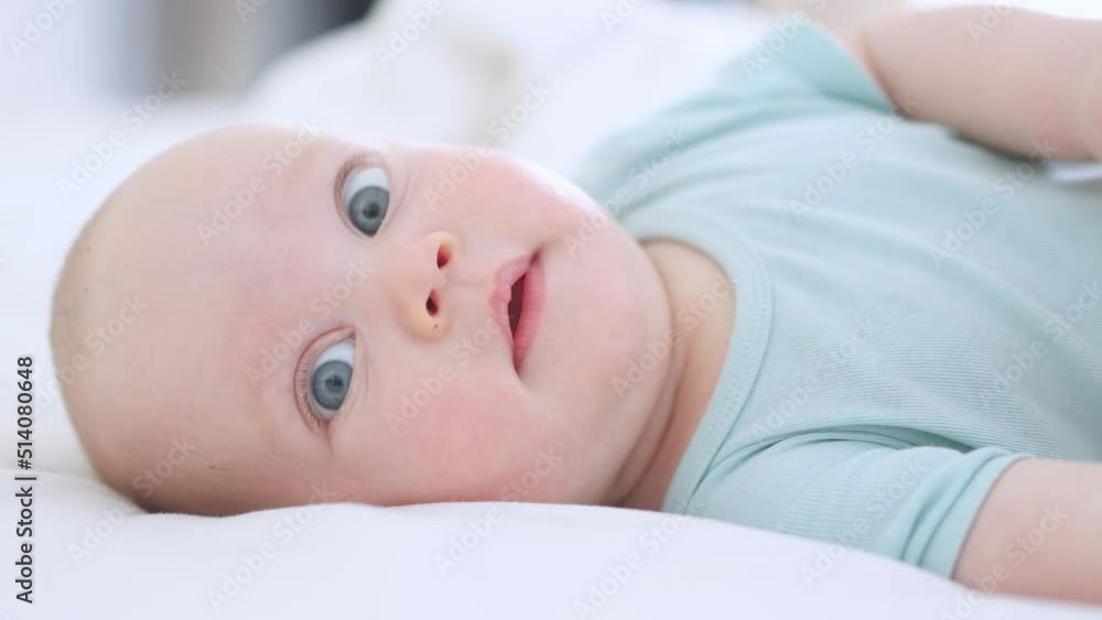 funny infant face portrait, blue eyes happy smiling baby child 3 months on white sheets. caucasian kid close-up portrait. happy little baby lying in bed looking into camera making funny grimaces. funny infant face portrait, blue eyes happy smiling baby child 3 months on white sheets. caucasian kid close-up portrait. happy little baby lying in bed looking into camera making funny grimaces.