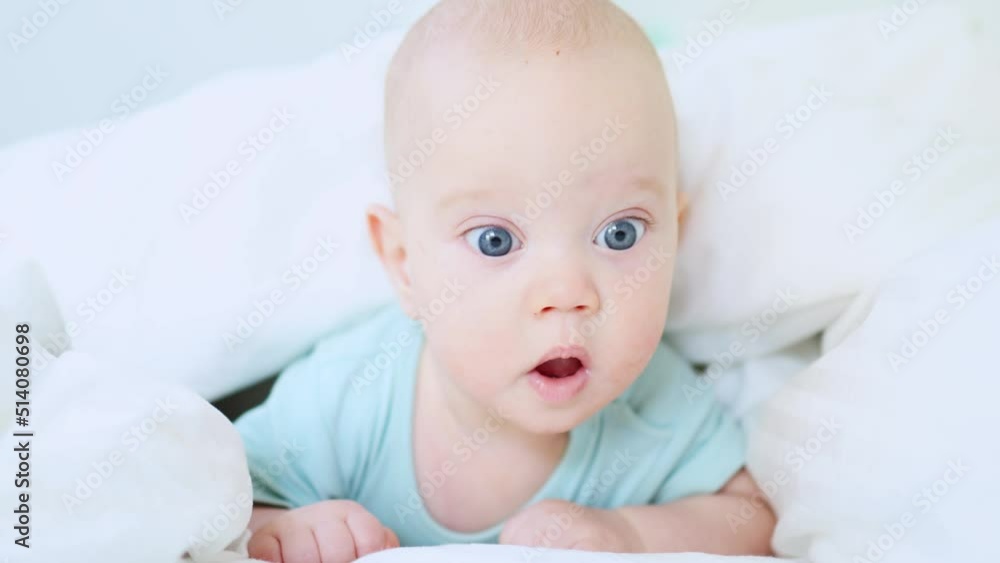 funny caucasian baby infant lying in white bed on stomach in the morning, happy 3 month child in blue clothes awake in sunny day bedroom, smiling portrait of blue-eyed kid in white bedsheets funny caucasian baby infant lying in white bed on stomach in the morning, happy 3 month child in blue clothes awake in sunny day bedroom, smiling portrait of blue-eyed kid in white bedsheets