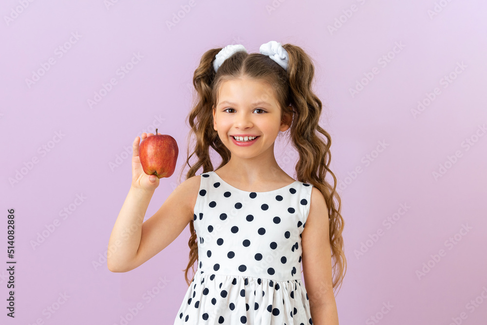 A little girl holds an apple and smiles. A beautiful child in a polka dot dress with curly hair holds a fruit on a pink isolated background. Vitamins for children. A little girl holds an apple and smiles. A beautiful child in a polka dot dress with curly hair holds a fruit on a pink isolated background. Vitamins for children.