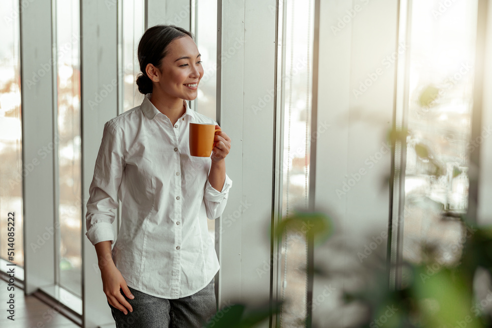 Fototapeta premium Portrait of asian business woman drinking coffee standing near windows at office. Blurred background