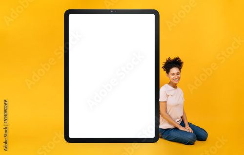 Full length photo of excited African American girl sits near an enlarged tablet with blank white mock-up screen for presentation or advertising, looks at camera, smiles, orange isolated background
