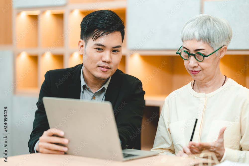 Young Asian business man in formal clothing showing something on digital tablet to senior female boss and manager wearing eyeglasses while in a meeting and training on technology Young Asian business man in formal clothing showing something on digital tablet to senior female boss and manager wearing eyeglasses while in a meeting and training on technology